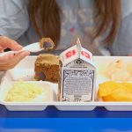Photo of a student eating a meal from a compartment tray at a school cafeteria. The tray contains a carton of milk, a slice of brown bread, scrambled eggs, and fruit pieces while the student holds a spoon with a bite of food.