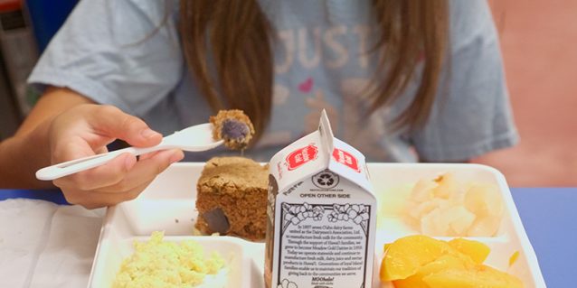 Photo of a student eating a meal from a compartment tray at a school cafeteria. The tray contains a carton of milk, a slice of brown bread, scrambled eggs, and fruit pieces while the student holds a spoon with a bite of food.